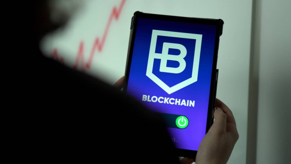 Person hält Tablet, dass auf dem Bildschirm das Wort Blockchain, darüber groß ein passendes Logo sowie darunter einen aktivierten Ein-Aus-Schalter anzeigt 