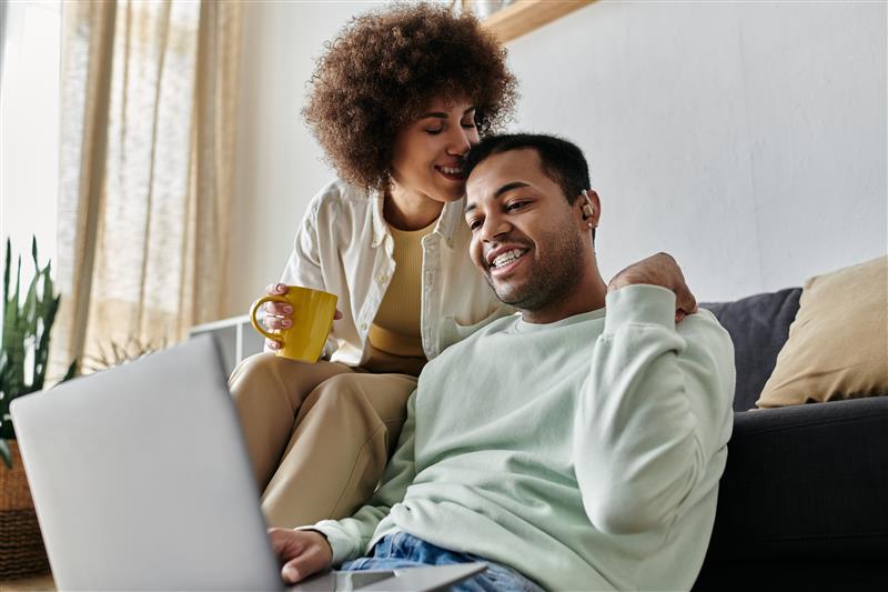 Eine liebevolle afroamerikanische Frau sitzt auf dem Sofa neben ihrem Mann, der ein Hörgerät trägt. Er schaut auf einen Laptop mit barrierefreien Inhalten.